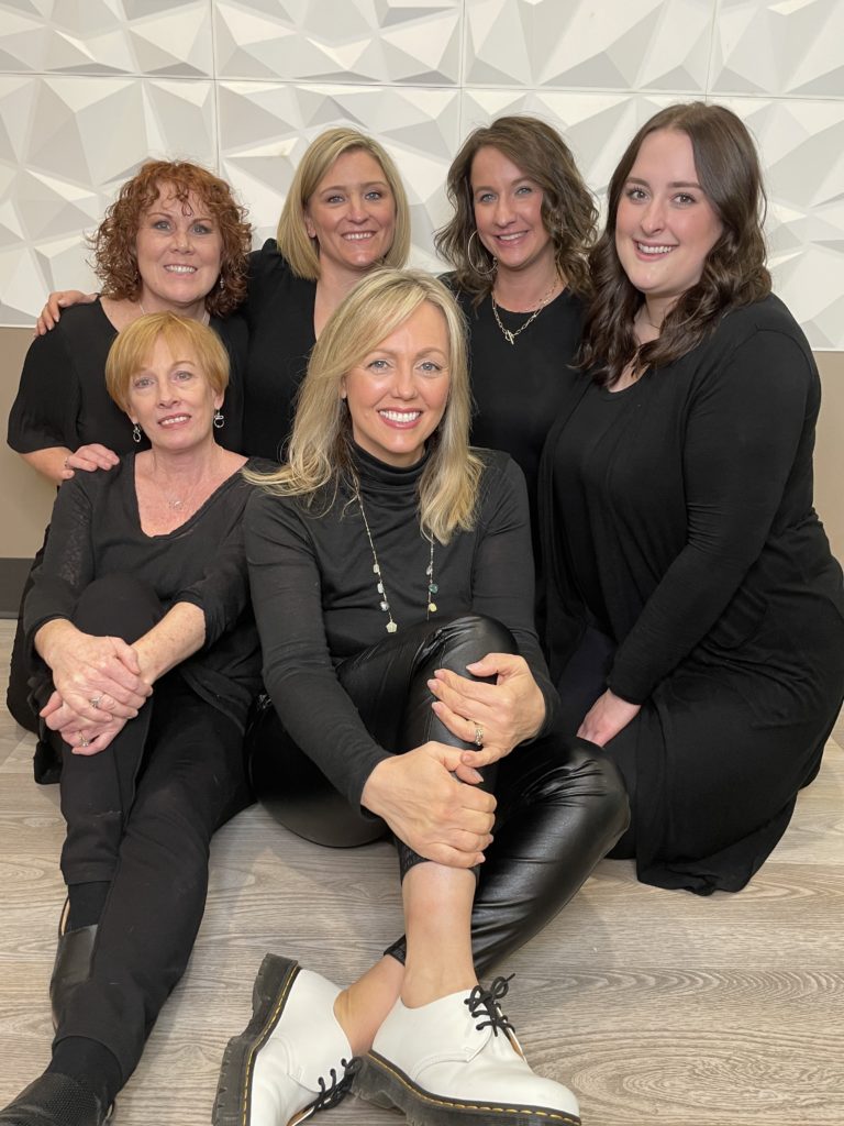 - Bella Salon in Madison, WI Six women dressed in black sit and kneel together on a wood floor, smiling at the camera against a white geometric wall, showing off stylish hair cuts and highlights. - Bella Salon in Madison, WI