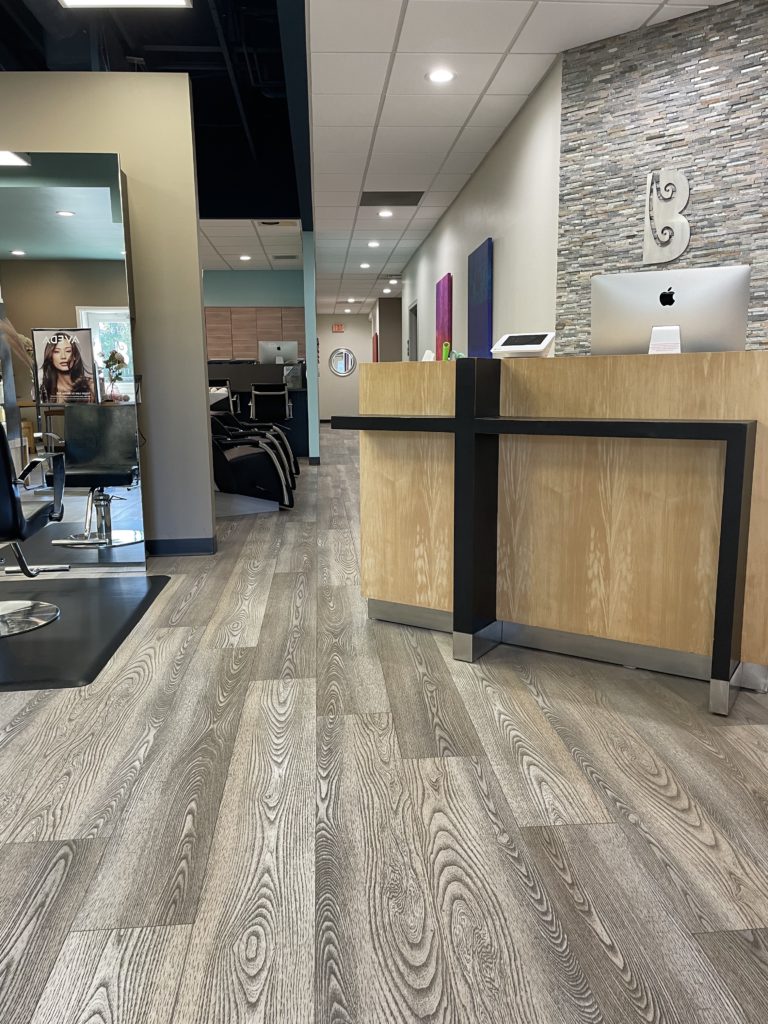 - Bella Salon in Madison, WI Modern hair salon interior with wood-look flooring, a wooden reception desk with an iMac, salon chairs, mirrors, and decorative wall accents—perfect for expert Hair Cuts and Plant Based Hair Care. - Bella Salon in Madison, WI