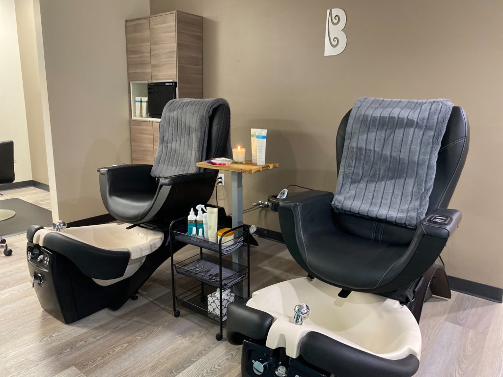 - Bella Salon in Madison, WI Two black pedicure chairs with foot basins, each covered with a gray towel, beside a small cart with nail supplies in a clean, modern salon setting that also offers highlights and smoothing treatments. - Bella Salon in Madison, WI